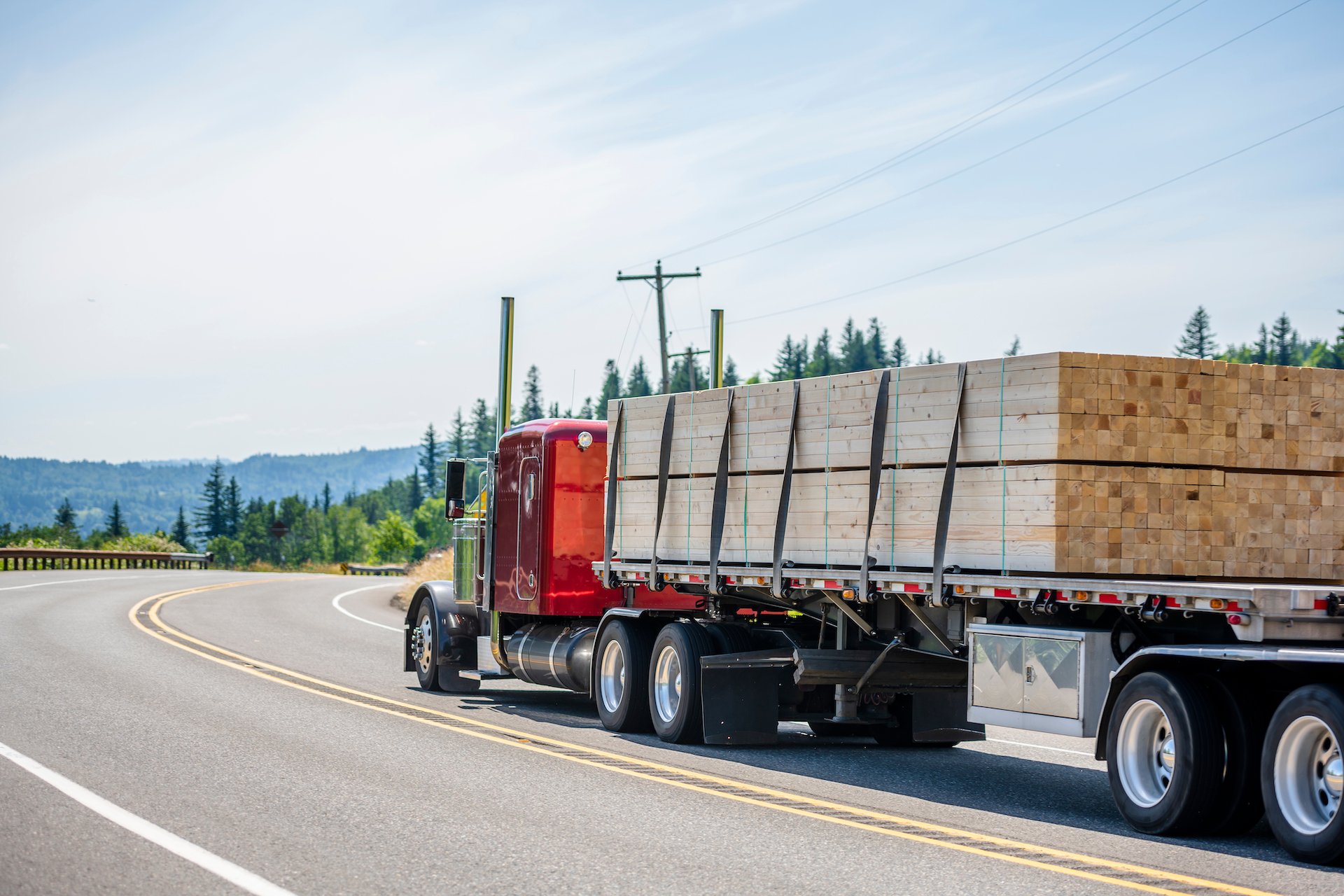 Big rig red classic powerful long haul semi truck with flat bed semi trailer transporting tightened industrial lumber boards running on the turning winding road with hills and trees in sunny day