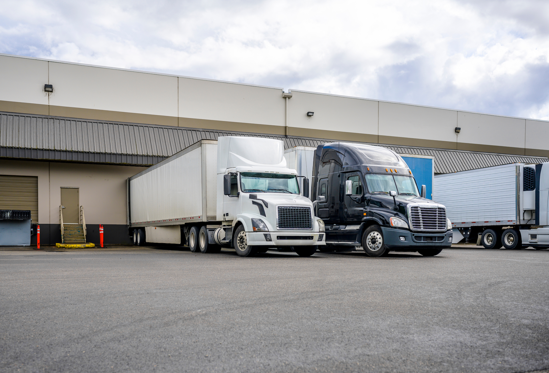 Big rig long haul industrial freight white semi truck loading commercial cargo to dry van semi trailer standing at the warehouse dock gate in row with another loaded semi trucks and semi trailers