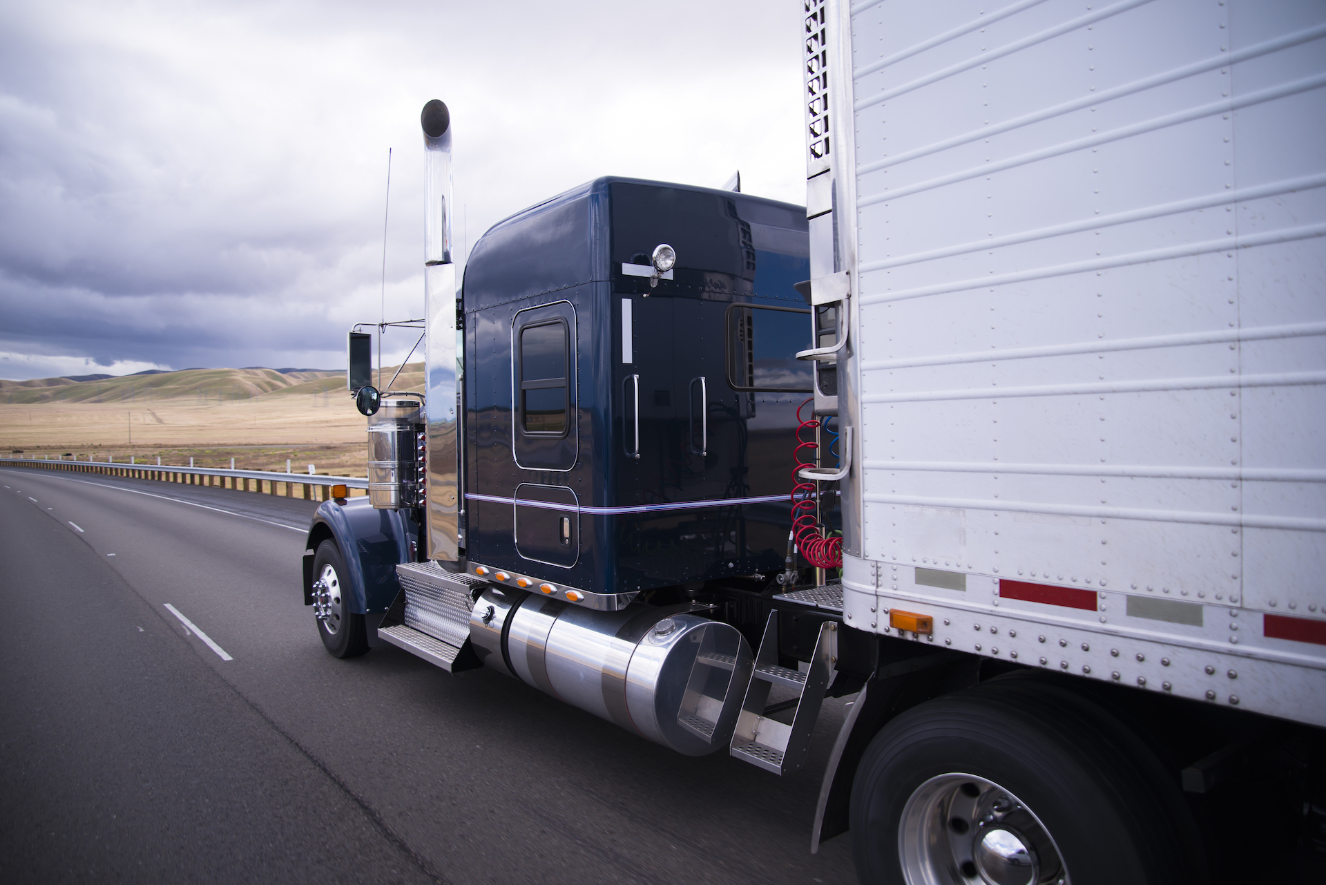 Classic bonneted American semi truck with chrome trim and a refrigerator trailer drive on the straight road in the California fields.