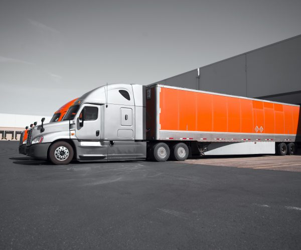 Two modern professional commercial semi trucks in gray and orange with dry van trailers are standing in the dock on territory of the transport warehouses for loading or unloading of commercial cargo